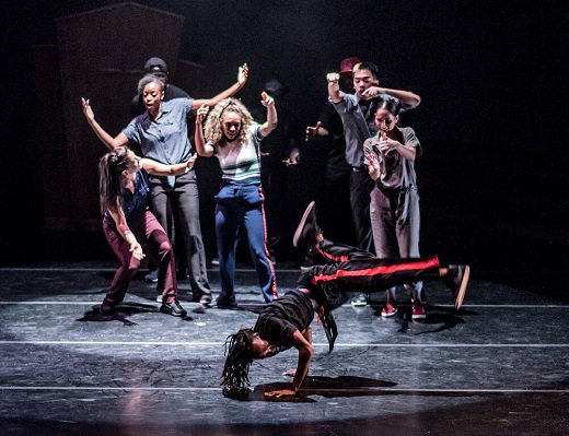 One of the Hood Lockers balances on his hands in a breakdance freeze, with head and legs hovering off the floor. Puremovement dancers surround him from behind, arms raised and pointing to him.